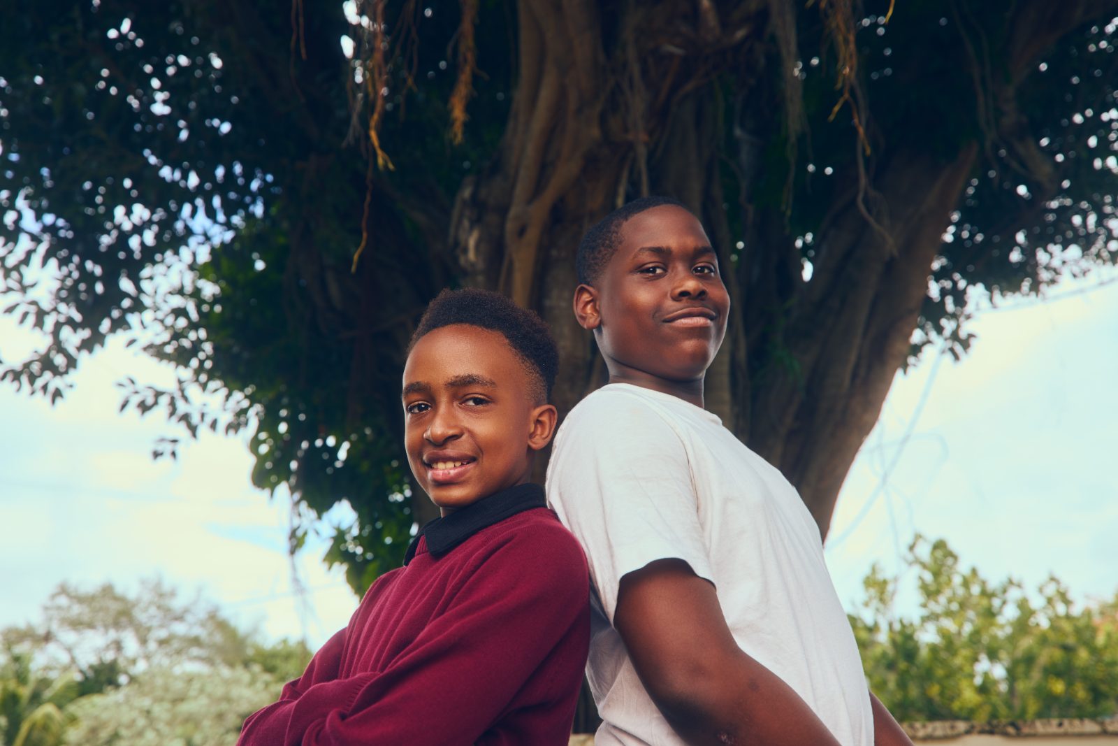 Two smiling students standing back to back under a large tree